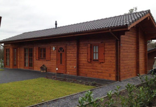 Een foto van een sfeervolle houten lodge met donker pannendak, gazon en grindpad, overdag genomen.