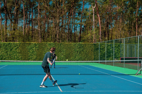 Une personne joue au tennis sur un court extérieur entouré de grands arbres et de verdure.