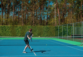 Una persona juega tenis en una cancha al aire libre rodeada de altos árboles y vegetación.