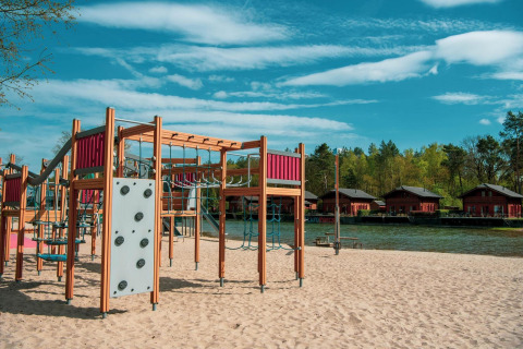 Parque infantil con estructuras para escalar sobre arena, junto a lago y cabañas de madera al fondo.