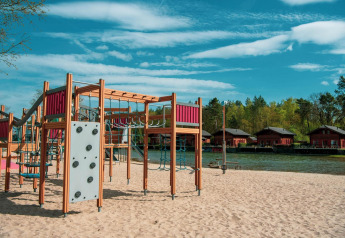 Speelplein op zand met klimrekken bij een waterplas, houten huisjes op de achtergrond van een lodge.