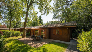 Vista soleada del lodge Boekhorst 6 de madera, rodeado de árboles, terraza exterior con mesa y sillas.