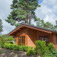 A cozy wooden lodge set among tall pine trees and greenery, featuring large windows and a garden view.