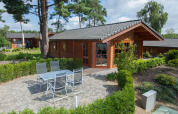 Photo of a cozy wooden lodge with a tiled patio, garden furniture, and lush green landscaping around.