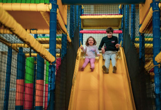 Dos niños bajan por un tobogán amarillo en una colorida zona de juegos interior en un glamping.