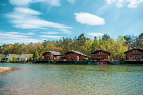 Scenic lakeside view of wooden cabins at EuroParcs glamping site, surrounded by forest and blue sky.
