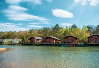 Scenic lakeside view of wooden cabins at EuroParcs glamping site, surrounded by forest and blue sky.