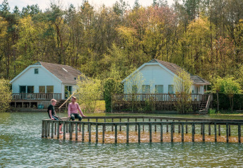 To personer sidder på en træbro ved søen foran hyggelige glamping-hytter omgivet af grøn skov.