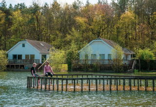 Dos personas se relajan en un muelle de madera junto al lago frente a cabañas glamping entre árboles.