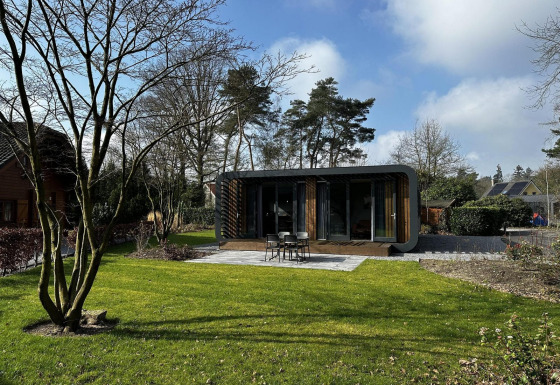 Moderne Modus Sauna 4 lodge met grote ramen in een groene tuin, omringd door bomen en open hemel.