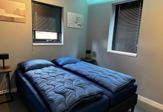 Moderne slaapkamer met twee eenpersoonsbedden, blauw beddengoed, donkere jaloezieën en minimalistisch interieur.
