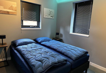 Moderne slaapkamer met twee eenpersoonsbedden, blauw beddengoed, donkere jaloezieën en minimalistisch interieur.