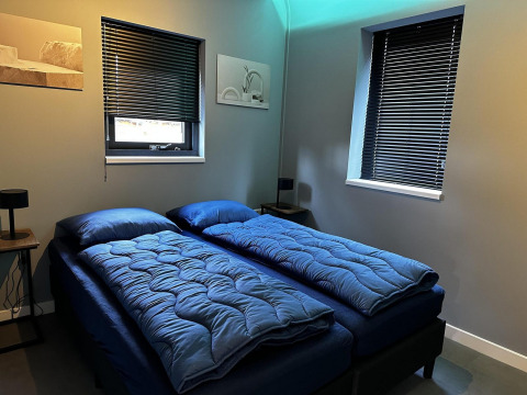 Modernes Schlafzimmer mit zwei Einzelbetten, blauer Bettwäsche, dunklen Jalousien und minimalistischer Deko.