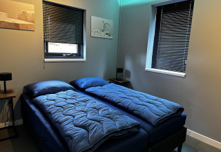 Modernes Schlafzimmer mit zwei Einzelbetten, blauer Bettwäsche, dunklen Jalousien und minimalistischer Deko.