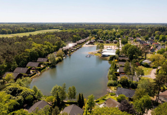 Vista aérea de un alojamiento glamping con cabañas junto al lago, zonas verdes y bosque alrededor.