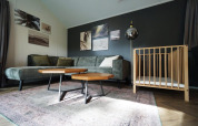 Stue i Baby Cottage-lodgen, med grøn sofa, træbabyseng, sofaborde og vægkunst på mørk væg.