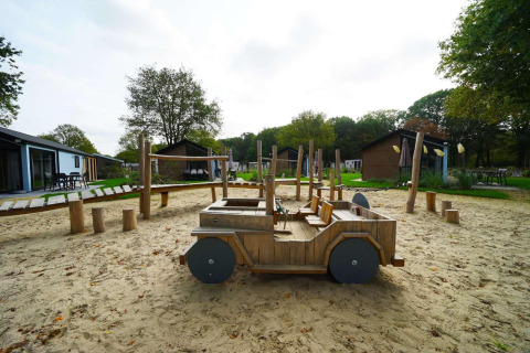 Parque infantil con coche de madera y arena en Baby Cottage lodge en Kaatsheuvel, Países Bajos, rodeado de cabañas.