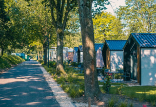 Una fila di lodge moderni si trova accanto a un sentiero alberato immerso nel verde in una giornata di sole.