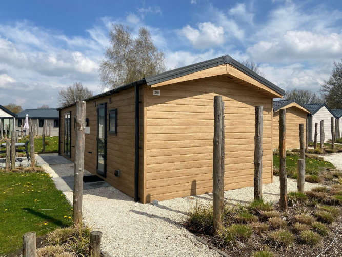 Modern Berkel lodge with wooden exterior, surrounded by small gardens under a blue sky with clouds.
