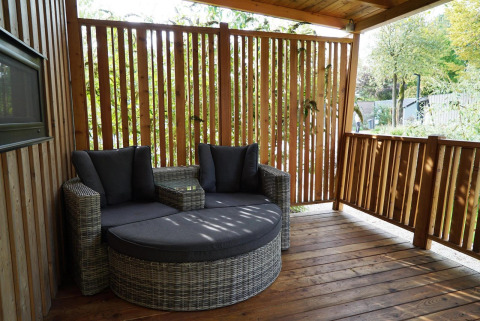 Covered patio with rattan sofa and cushions at Cottage at Kaatsheuvel, Netherlands, surrounded by wood.