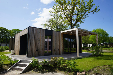 Lodge Domus moderna con paneles de madera, terraza y vegetación, rodeada de árboles y cielo claro.