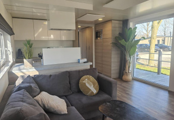 Moderne leefruimte in lodge Domus in Kaatsheuvel, Nederland, met grote ramen, grijze zetel en keukenhoek.