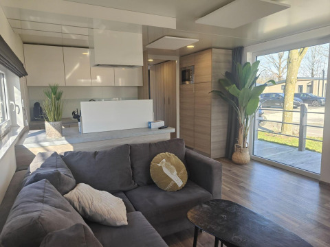 Modern living room in Domus lodge, Kaatsheuvel, Netherlands, with large windows, gray sofa, and kitchen area.