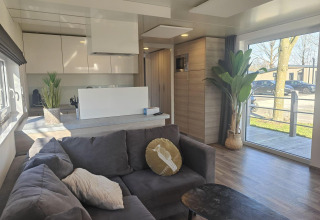 Modern living room in Domus lodge, Kaatsheuvel, Netherlands, with large windows, gray sofa, and kitchen area.