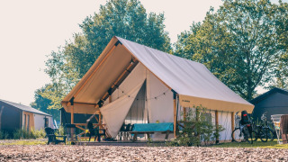 Tienda safari de glamping bajo el sol, con muebles exteriores, bicicletas y árboles verdes alrededor.