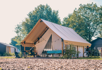 En moderne glamping-safariteltsplads i solskin med cykler, havemøbler og grønne træer i baggrunden.