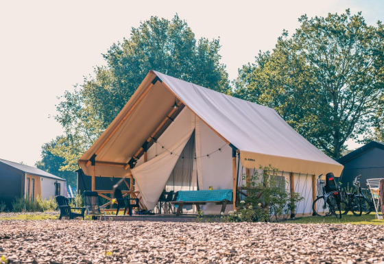 Moderne glamping safaritent op een zonnige camping, met buitenzitjes, fietsen en groene bomen.