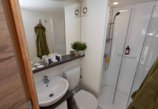 Baño moderno dentro de una tienda glamping con ducha, lavabo, inodoro y toallas verdes colgadas.