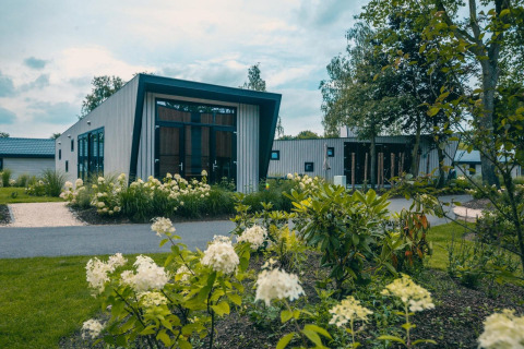 Lodge Just Nature moderne avec de grandes fenêtres, entouré de jardins luxuriants et de plantes fleuries.