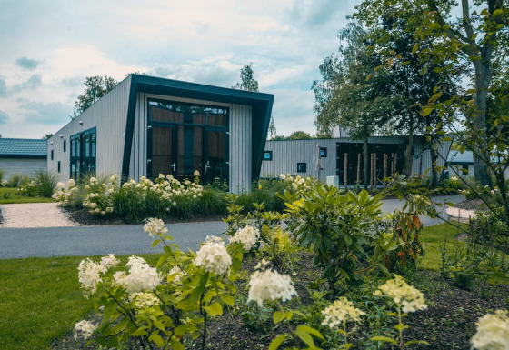 Lodge Just Nature moderne avec de grandes fenêtres, entouré de jardins luxuriants et de plantes fleuries.