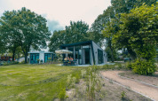 En moderne hytte Just Nature i Kaatsheuvel, Holland, med folk der sidder udendørs omgivet af grønne træer.