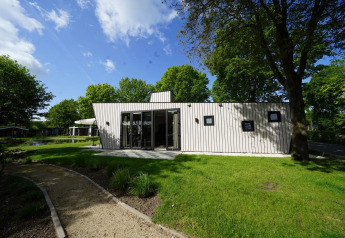 Lille hus Just Nature i Kaatsheuvel, Holland, omgivet af grønt græs og træer under en blå himmel.