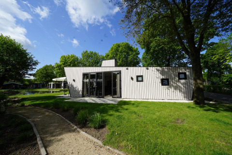 Lille hus Just Nature i Kaatsheuvel, Holland, omgivet af grønt græs og træer under en blå himmel.