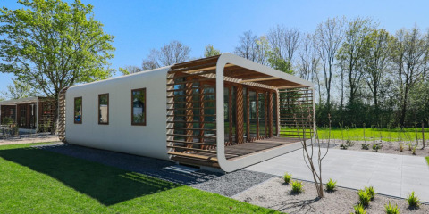 Moderne lodge met afgeronde hoeken en houten latten, omringd door gras en bomen op een zonnige dag.