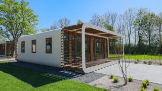 Modern lodge with curved edges and wooden slats, surrounded by green grass and trees on a sunny day.