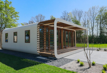 Lodge moderne aux bords arrondis et lattes en bois, entouré de pelouse verte et d’arbres par une journée ensoleillée.