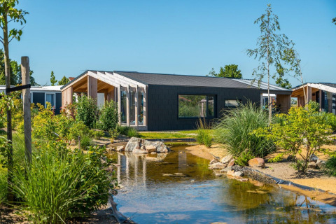Moderne Panorama Cottage 4-lodge med stor vindue, have og dam under en klar, blå himmel.