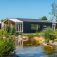 Moderne Panorama Cottage 4-lodge med stor vindue, have og dam under en klar, blå himmel.