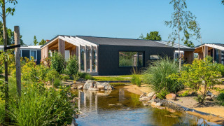 Panorama Cottage 4: lodge moderno con ampia finestra, giardino e stagno sotto cielo azzurro.