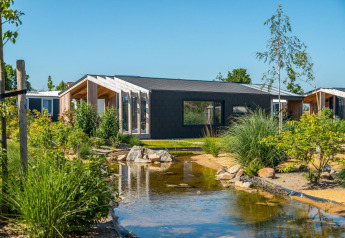 Moderne Panorama Cottage 4-lodge med stor vindue, have og dam under en klar, blå himmel.