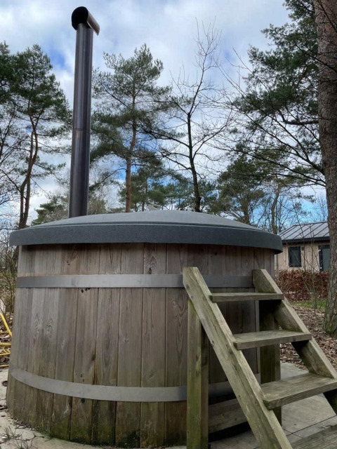 Bañera de hidromasaje de madera al aire libre con chimenea y escalera junto a una cabaña rodeada de árboles.