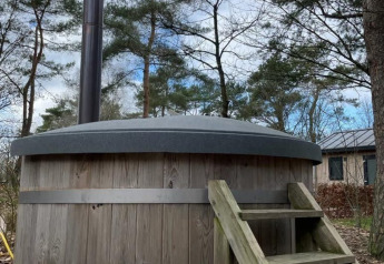 Outdoor wooden hot tub with a chimney and steps, located next to a lodge and surrounded by tall trees.
