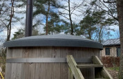 Buiten houten hottub met schoorsteen en trap, naast een lodge en omringd door hoge bomen.