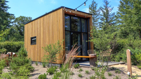 Casa piccola moderna chiamata Tiny House 2 con rivestimento in legno, grandi finestre e giardino verde intorno.