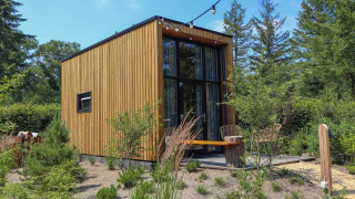 Casa piccola moderna chiamata Tiny House 2 con rivestimento in legno, grandi finestre e giardino verde intorno.