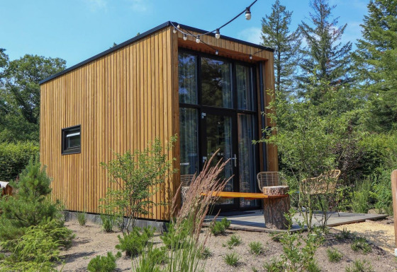 Casa piccola moderna chiamata Tiny House 2 con rivestimento in legno, grandi finestre e giardino verde intorno.